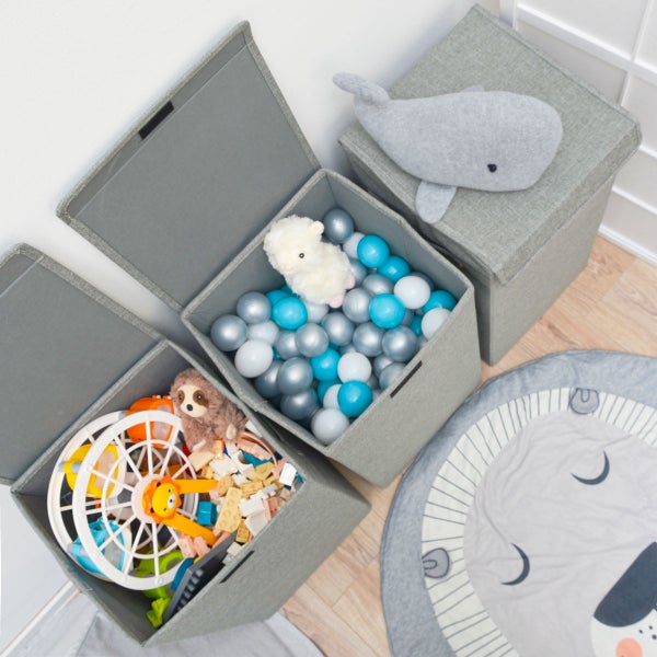 Drei graue Aufbewahrungsboxen aus Stoff in einem hellen Kinderzimmer. Eine ist geschlossen, zwei sind mit Spielzeug und Bällen gefüllt.