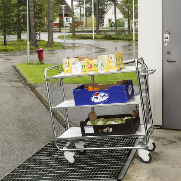 Mobiler Etagenwagen aus Metall mit drei weißen Ebenen und Drahtgitterwänden, beladen mit Obst- und Gemüsekisten im Außenbereich.