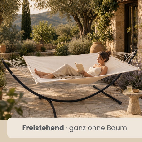 Freistehende Hängematte mit Metallgestell auf einer hellen Terrasse, eine Frau liest darin ein Buch.