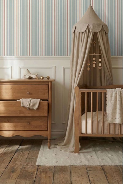 Ein Kinderzimmer mit gestreifter Tapete, einem Babybett mit Himmel aus Stoff, einer Kommode aus Holz und einem Mobile über dem Bett.