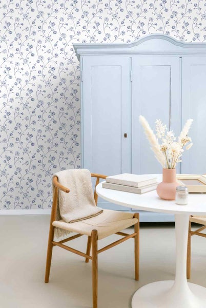 Florale Tapete mit blauen Blumenranken in einem hellen Zimmer mit einem Schrank, einem Holzstuhl und einem runden Tisch.