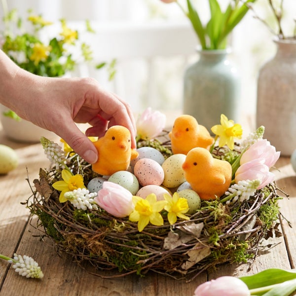 Eine Hand platziert ein Osterküken aus beflocktem Material in einem Nest mit Ostereiern und Frühlingsblumen wie Narzissen und Tulpen.