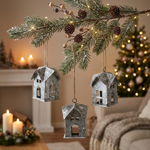 Drei hängende Weihnachtsdekorationshäuser aus Metall an einem künstlichen Tannenzweig mit Zapfen und Lichterkette in einem weihnachtlichen Raum.