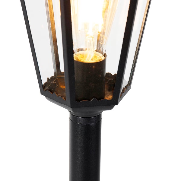 Nahaufnahme einer klassischen Außenleuchte mit schwarzem Metallrahmen, klaren Glasscheiben und einem leuchtenden Filament-Leuchtmittel.