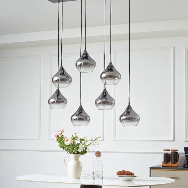 Moderne siebenflammige Pendelleuchte mit tropfenförmigen Glasschirmen über einem weißen Esstisch in einem hellen Esszimmer mit Wandpaneelen.