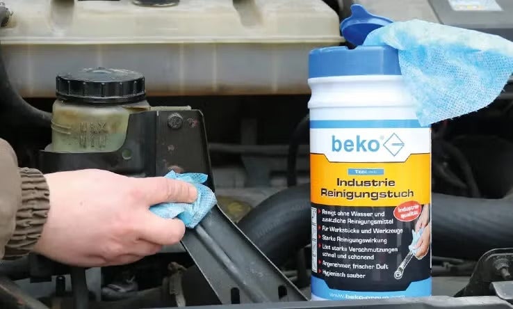 Eine Hand reinigt mit einem beko Industriereinigungstuch ein Bauteil im Motorraum eines Autos.