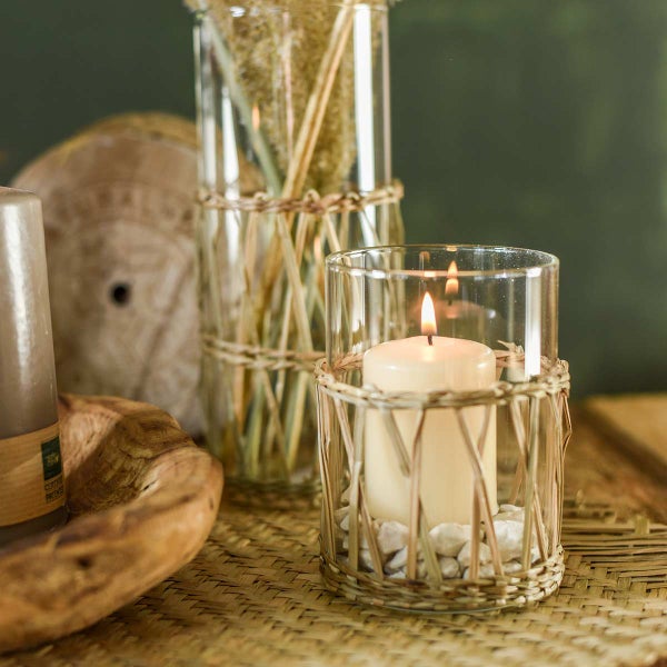 Windlicht aus Glas mit natürlichem Geflecht, darin eine brennende Stumpenkerze auf weißen Steinen.