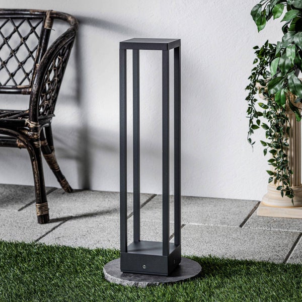 Moderne schwarze Außenstehleuchte mit offenem rechteckigem Rahmen auf einer hellen Terrasse mit Kunstrasen, Steinplatten und einem Rattansessel.