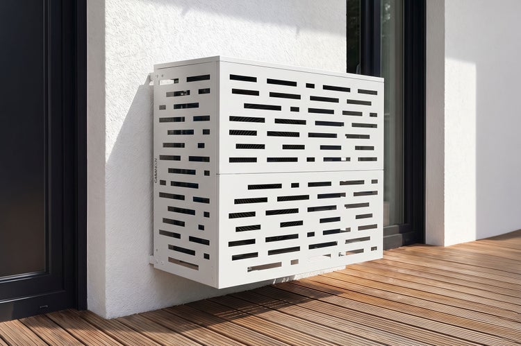 Wandmontierte Verkleidung für Klimaanlagen aus weißem Metall mit Schlitzmuster, montiert an einer Hausfassade über einer Holzterrasse.