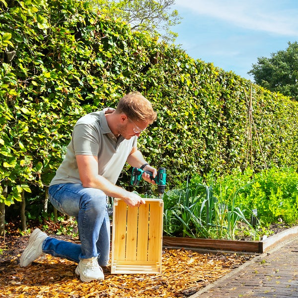 Ein Mann mit Schutzbrille benutzt einen Akkuschrauber an einer Holzkiste im Garten neben einem Hochbeet und einer Hecke.
