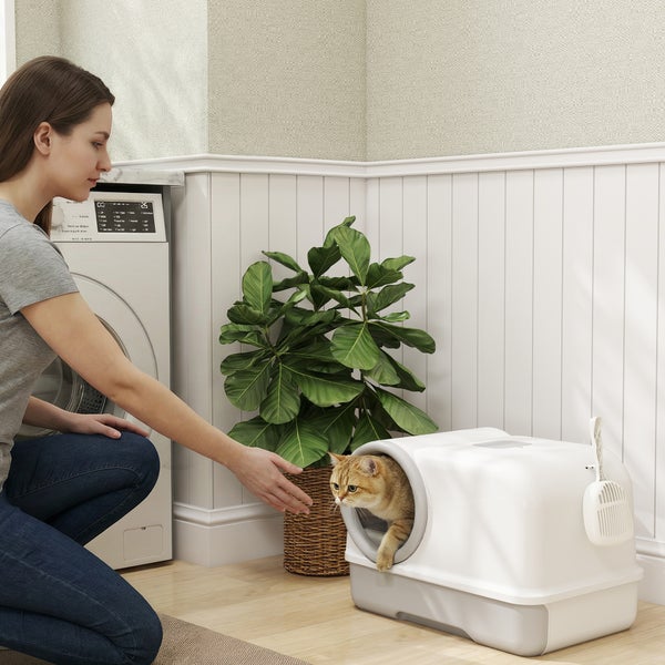 Frau kniet neben einer Haubentoilette für Katzen mit Schaufel, während eine Katze heraustritt. Heller Hauswirtschaftsraum mit Waschmaschine.