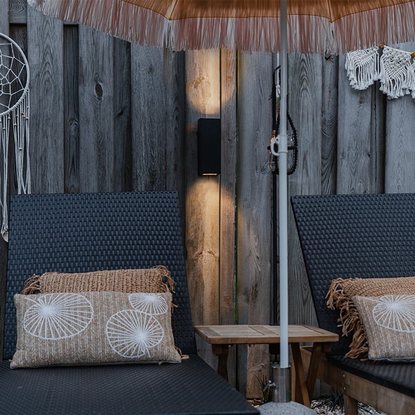 Schwarze Außenwandleuchte mit Licht nach oben und unten an einer Holzwand hinter Sonnenliegen auf einer Terrasse.