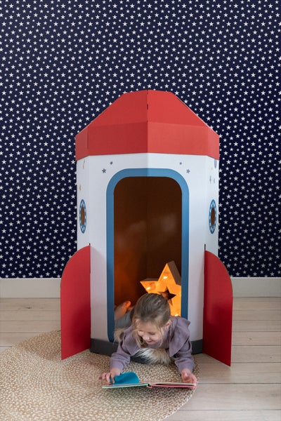 Mädchen liest Buch in einem Raketenspielhaus auf einem Teppich vor einer Wand mit Sternenmuster.