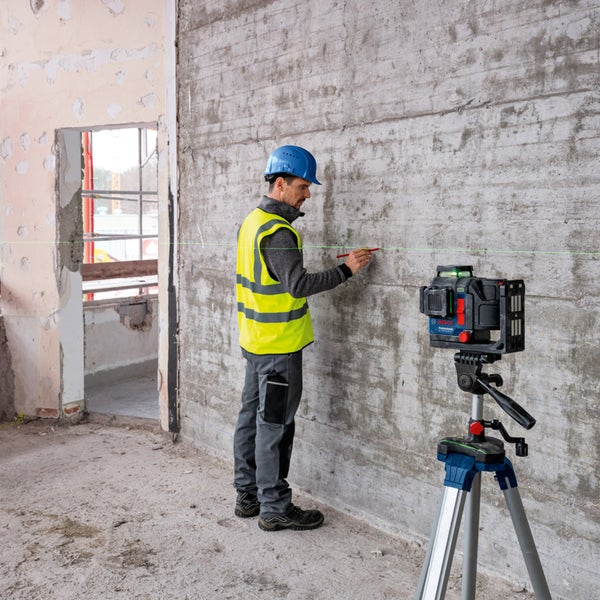 Ein Bauarbeiter markiert eine grüne Laserlinie an einer Wand mit einem Bleistift, wobei ein Linienlaser auf einem Stativ steht.