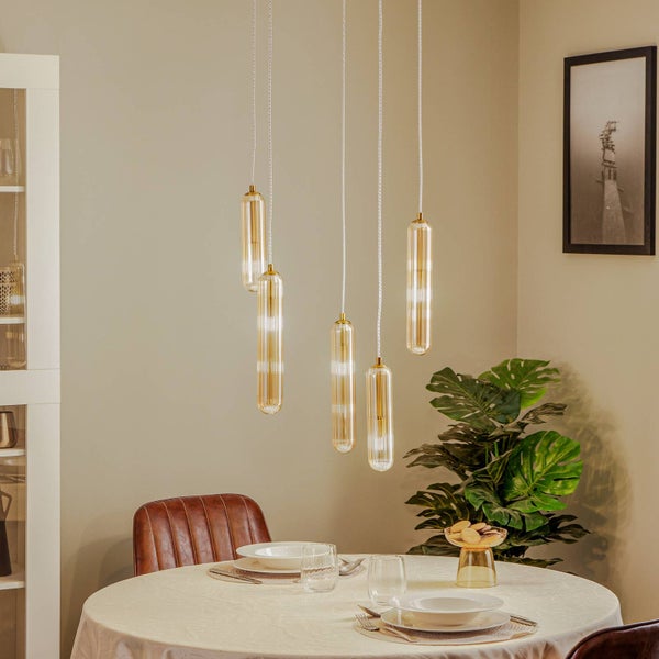 Moderne mehrflammige Pendelleuchte mit fünf langen, zylindrischen Glasschirmen in Gold über einem runden Esstisch in einem hellen Raum.