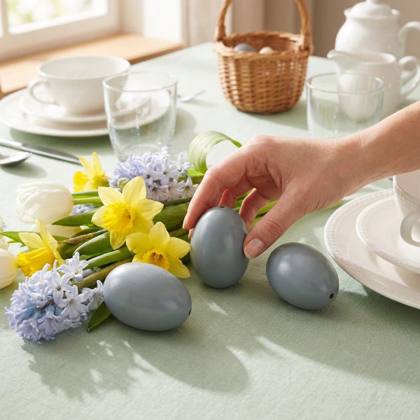 Hand platziert dekorative Ostereier neben Osterglocken und Hyazinthen auf einem gedeckten Tisch als frühlingshafte Tischdekoration.