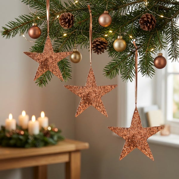 Drei hängende Weihnachtssterne mit strukturierter Oberfläche an einem Tannenbaumzweig mit Weihnachtskugeln, Tannenzapfen und Lichterkette.