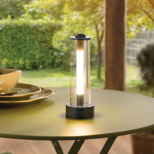 Tragbare LED-Außenleuchte mit transparentem Zylinder und schwarzem Sockel auf einem Metalltisch in einem hellen Garten.