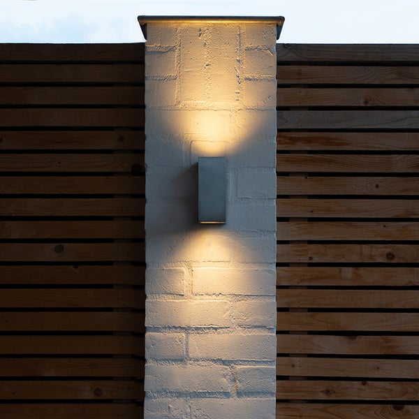 Rechteckige Außenwandleuchte an einem gemauerten Pfeiler mit Lichtaustritt nach oben und unten neben einem Holzzaun.