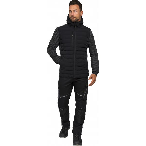 Mann in schwarzer Hybrid-Arbeitsjacke mit Kapuze und gestreiften Ärmeln, schwarzer Arbeitshose mit Taschen und dunklen Arbeitsschuhen.