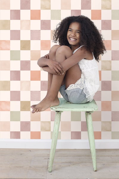 Mädchen mit lockigem Haar sitzt auf einem Hocker vor einer Wand mit quadratischem Muster.