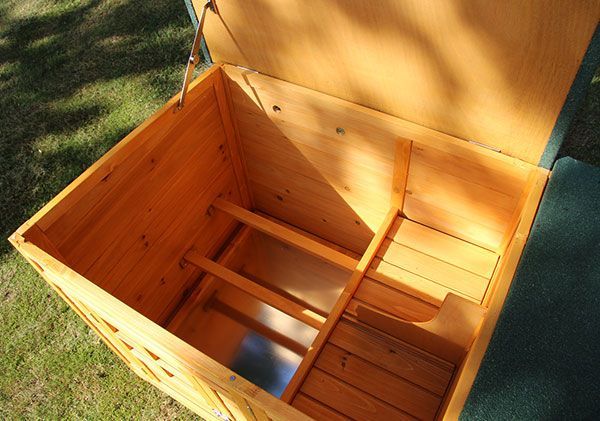 Innenansicht eines geöffneten Kleintierstalls aus Holz mit Nestbereich, Sitzstangen und einer Metallwanne.