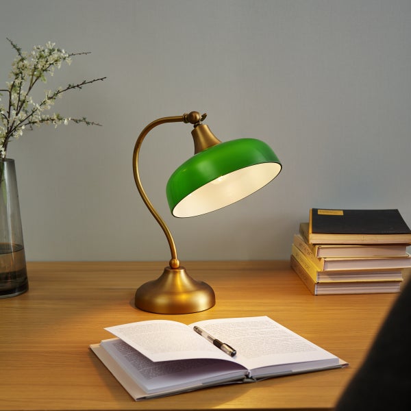 Klassische Schreibtischleuchte aus Messing mit grünem Metallschirm auf einem Holztisch neben Büchern und einem Notizheft.