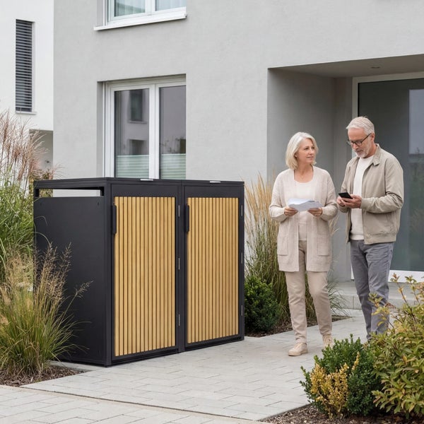 Moderne Zweier-Mülltonnenbox aus dunklem Metall mit hellen Holzlamellen im Außenbereich vor einem Wohnhaus.