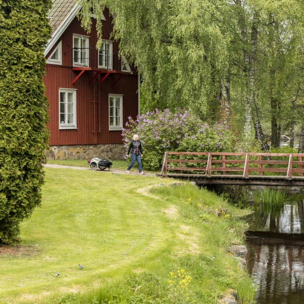 Szene mit einem Garten, einem Bach mit einer Holzbrücke und einer Frau, die einen Handwagen zieht