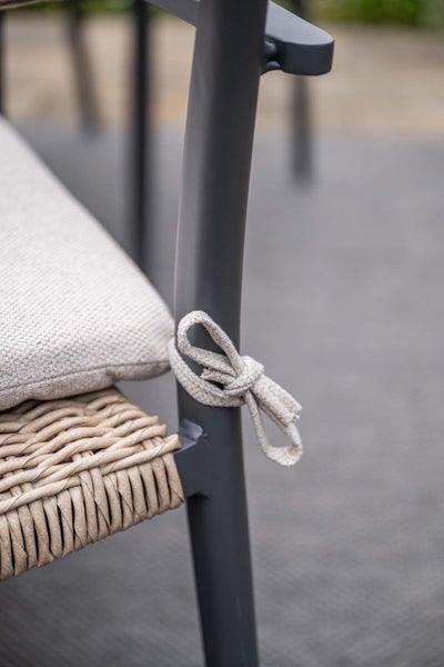 Detailaufnahme eines Gartenstuhls mit Sitzkissen, befestigt mit einer Schleife am grauen Metallgestell und beigen Geflecht.