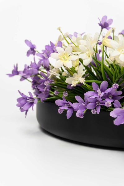 Künstliche Blumenanordnung mit lila und weißen Blüten in einer flachen schwarzen Schale.