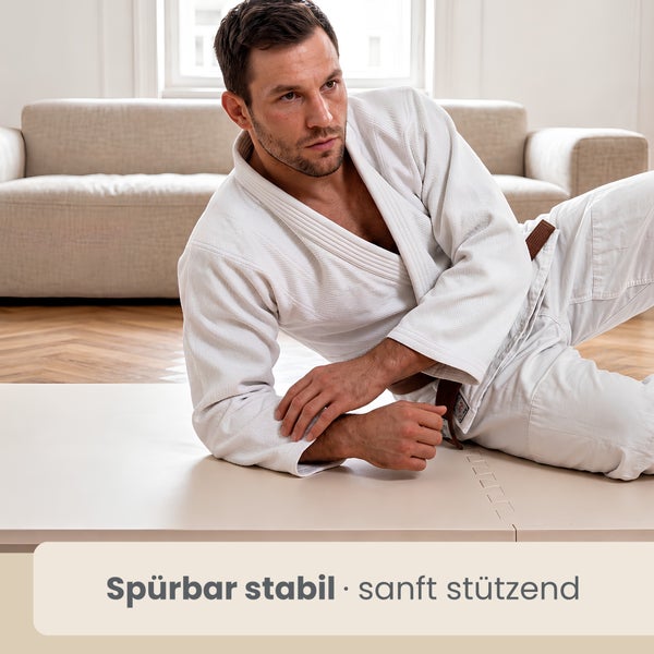 Mann im weißen Kampfsportanzug auf einer hellen Puzzle-Sportmatte im Wohnzimmer.