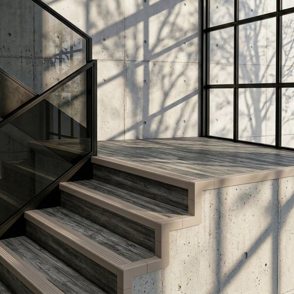 Moderne Treppe mit Bodenbelag in Holzoptik, Treppenkantenprofilen und Glasgeländer vor einer Sichtbetonwand.
