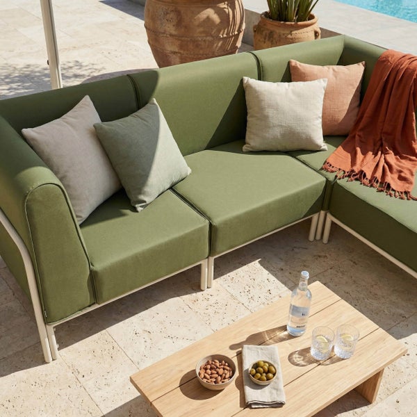 Modernes Garten-Lounge-Set mit grünem Ecksofa, hellen Kissen und einem Couchtisch aus Holz auf einer hellen Steinterrasse.