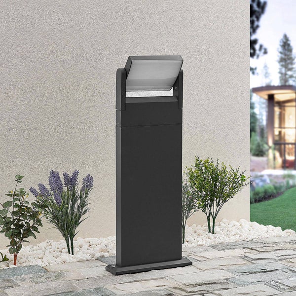 Moderne Außen-Sockelleuchte in Anthrazit mit schwenkbarem Kopf auf einer Terrasse mit Pflanzen und weißem Kies vor einer hellen Wand.