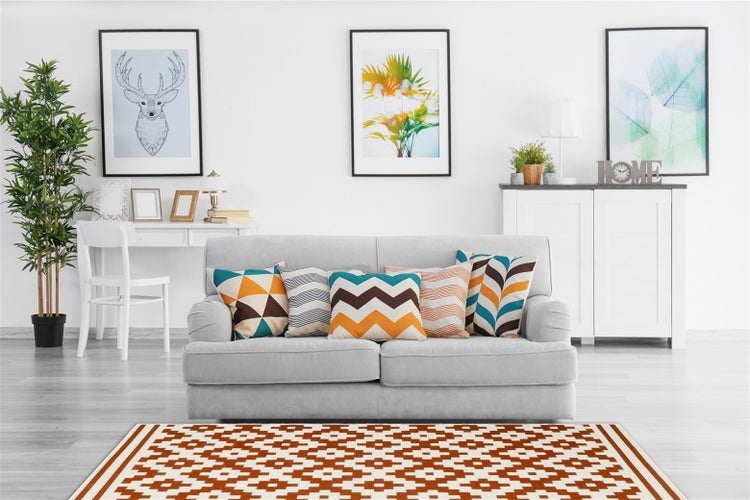 Wohnzimmer mit grauem Sofa, gemusterten Kissen, weißem Sideboard, Wandbildern, Zimmerpflanze und einem geometrisch gemusterten Teppich auf dem Boden.