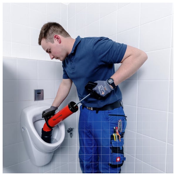 Ein Handwerker reinigt ein weißes Urinal in einem hellen, gefliesten Badezimmer mit einer roten Sanpro Saug-Druck-Rohrreinigungspumpe.