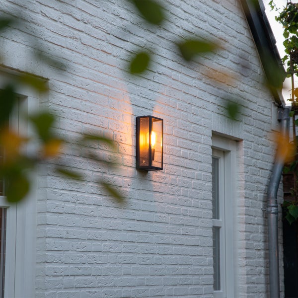 Moderne Außenwandleuchte in Laternenform aus schwarzem Metall und Glas, montiert an einer weißen Ziegelwand, strahlt warmes Licht aus.