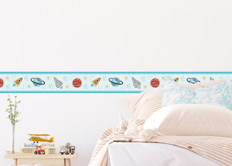 Selbstklebende Wandbordüre mit Weltraum-Motiven wie Raketen und Planeten in einem hellen Kinderzimmer über einem Bett.