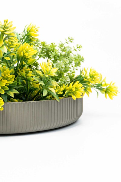 Künstliche Pflanze mit gelben Blüten in einer runden, grau geriffelten dekorativen Schale.