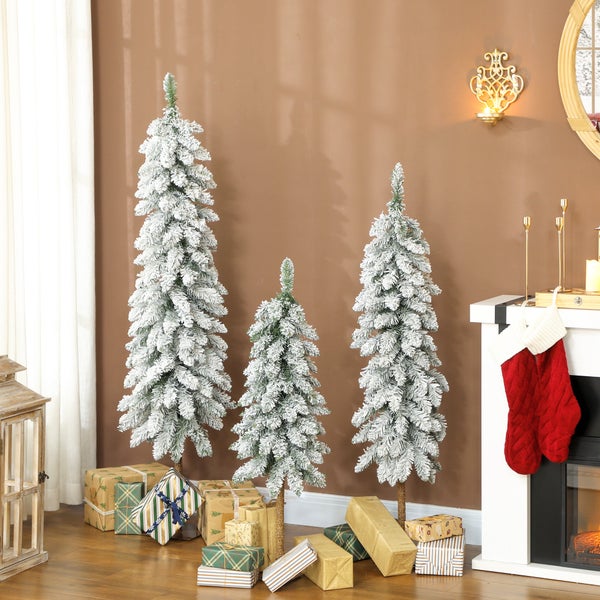 Drei schmale künstliche Weihnachtsbäume in verschiedenen Höhen mit Kunstschnee, umgeben von Geschenken neben einem Kamin.