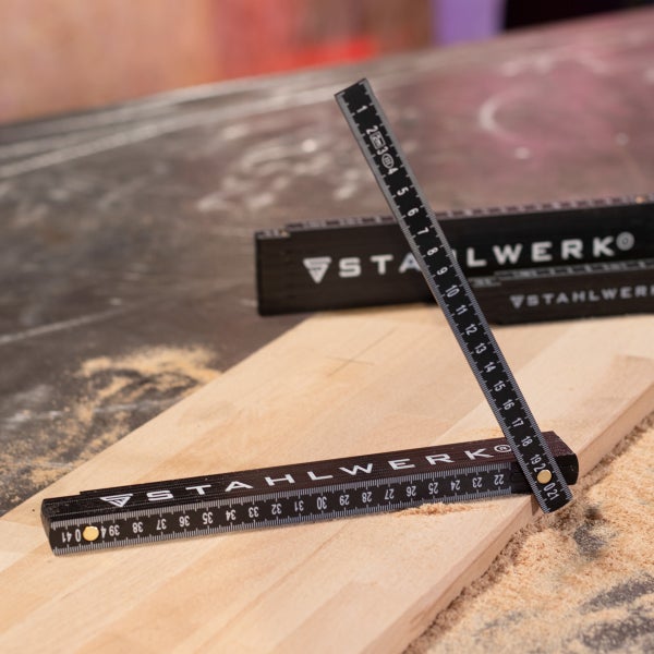 Schwarzer Gliedermaßstab von Stahlwerk, teilweise ausgeklappt auf einem Holzbrett mit Sägespänen.