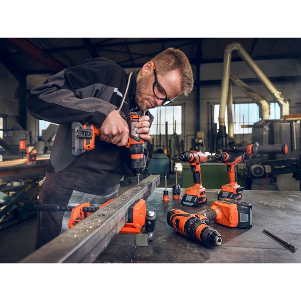 Ein Mann bohrt mit einem Akkubohrer in ein Metallstück, mehrere Bohrmaschinen liegen auf einem Tisch.