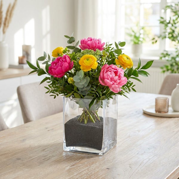 Blumenstrauß aus Pfingstrosen und Ranunkeln in einer quadratischen Glasvase mit schwarzem Dekosand auf einem Holztisch.