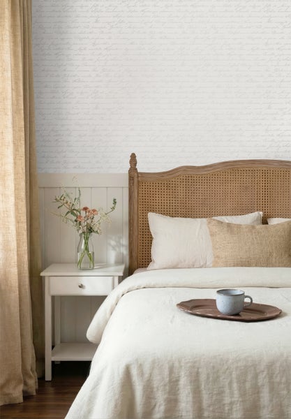 Schlafzimmer mit Bett inklusive Rattankopfteil, weißem Nachttisch und Vliestapete mit Handschrift-Muster über weißer Wandvertäfelung.