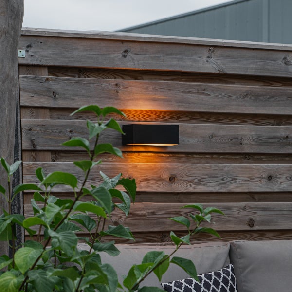 Rechteckige schwarze Außenwandleuchte mit Up-and-Down-Lichteffekt an einem Holzzaun im Garten.