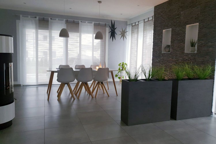Helle moderne Esszimmer-Szene mit Tisch, weißen Stühlen, Fliesenboden, Steinwand und großen Pflanzkübeln mit Ziergras im Vordergrund.