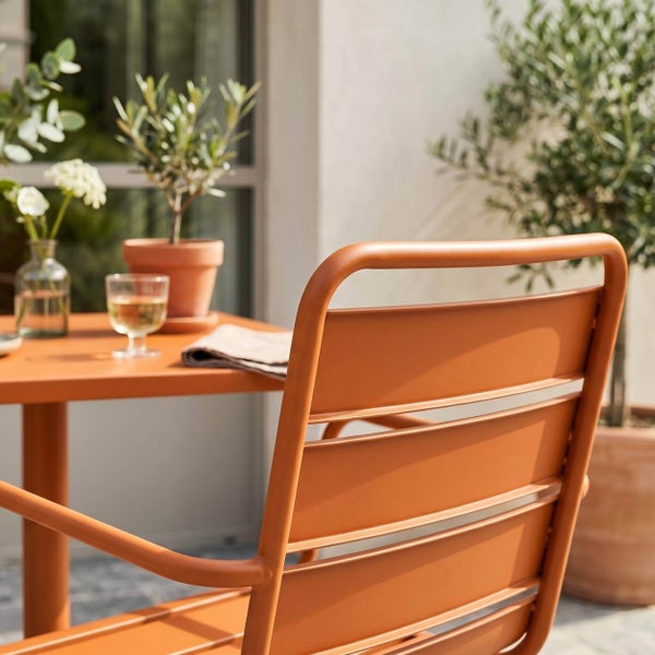 Detailaufnahme eines Metall-Gartenstuhls mit Lamellen-Rückenlehne an einem passenden Tisch im Außenbereich, dekoriert mit Pflanzen und einem Glas.