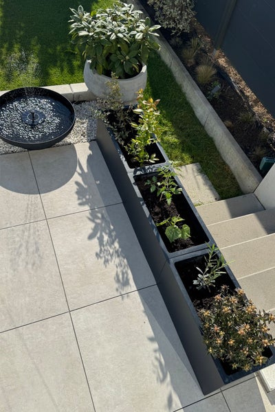Moderner Garten mit rechteckigem Pflanzkasten, rundem Wasserspiel und hellen Steinplatten auf der Terrasse.