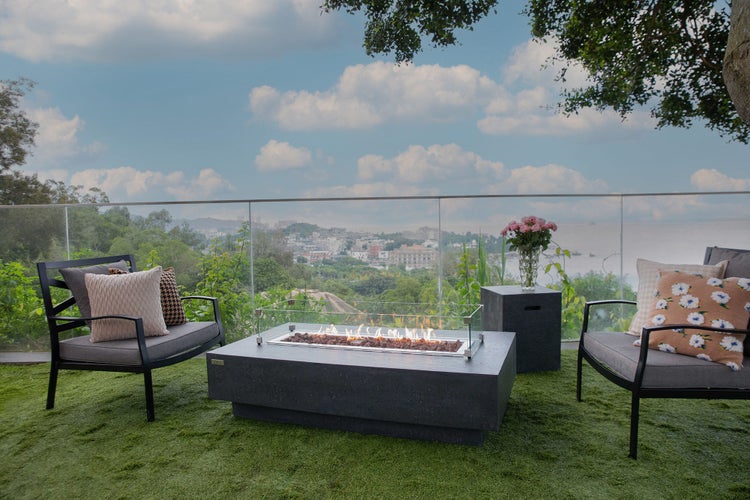 Moderner, heller Außenbereich mit einer rechteckigen Gas-Feuerstelle aus Beton, Metallsesseln mit Kissen und Glasgeländer vor Stadtlandschaft.
